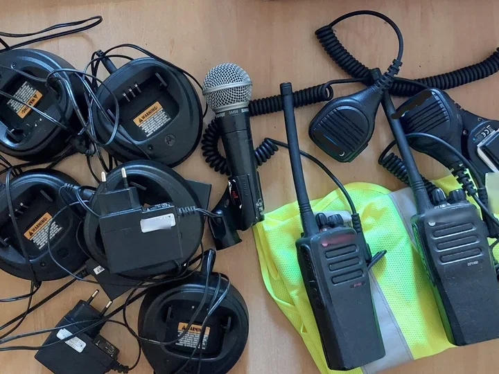 Na drewnianym blacie leżą krótkofalówki, ładowarki, mikrofon i odblaskowa kamizelka.