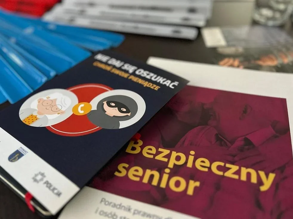 Na stole leżą materiały edukacyjne dla seniorów, w tym poradnik „Bezpieczny senior” i broszura „Nie daj się oszukać” z logiem policji.