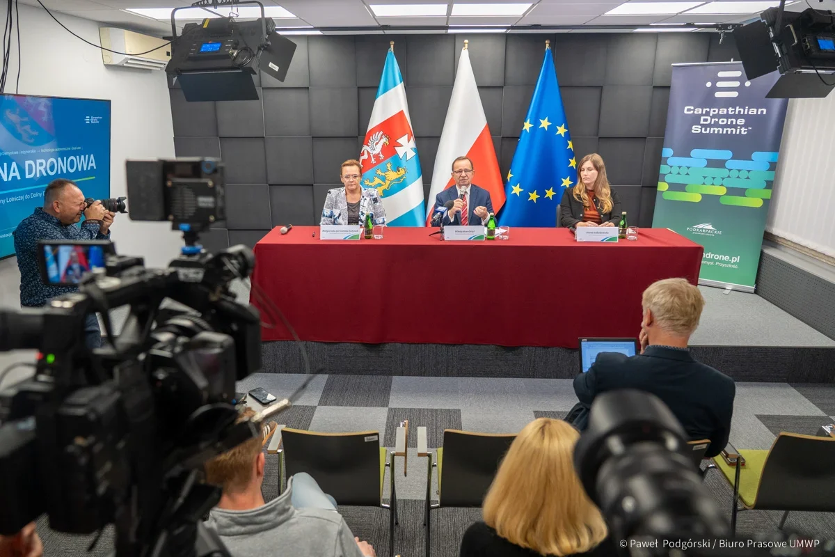 Trzy osoby siedzą za czerwonym stołem podczas konferencji prasowej z flagami w tle, na sali z kamerzystami i fotoreporterami.