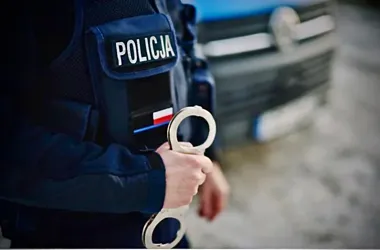 Policjant w mundurze z napisem POLICJA trzyma w ręku kajdanki, w tle widać policyjny radiowóz.