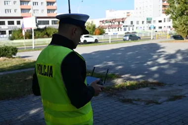 Policjant w żółtej kamizelce z napisem POLICJA obsługuje drona w miejskiej okolicy.