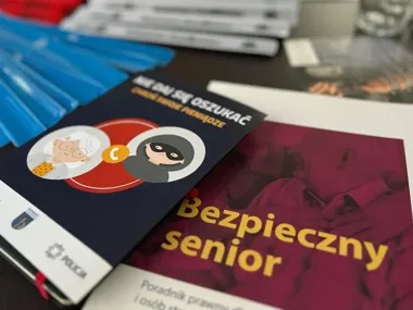 Na stole leżą materiały edukacyjne dla seniorów, w tym poradnik „Bezpieczny senior” i broszura „Nie daj się oszukać” z logiem policji.