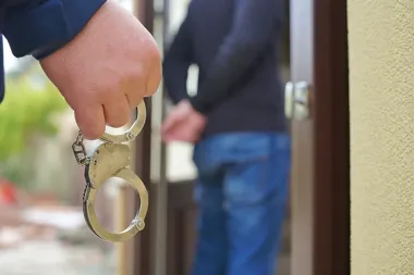 Funkcjonariusz trzyma kajdanki przy otwartych drzwiach, w tle stoi mężczyzna w dłoniach założonych za plecy.